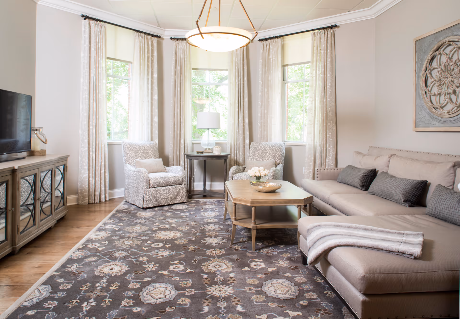 A bright and cozy living room with a large beige sectional sofa adorned with gray pillows and a folded blanket. Two patterned armchairs flank a round wooden side table with a lamp, positioned in front of three windows with sheer curtains. A wooden coffee table with decorative items sits on a large patterned area rug. A flat-screen TV is on a wooden cabinet to the left, and a decorative wall piece hangs above the sofa.
