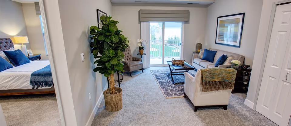 A cozy living room with a beige sofa, two armchairs, a coffee table on a patterned rug, and a large potted plant near the wall. The room has a sliding glass door leading to an outdoor area. To the left, a bedroom with a bed, nightstand, and lamp is partially visible.