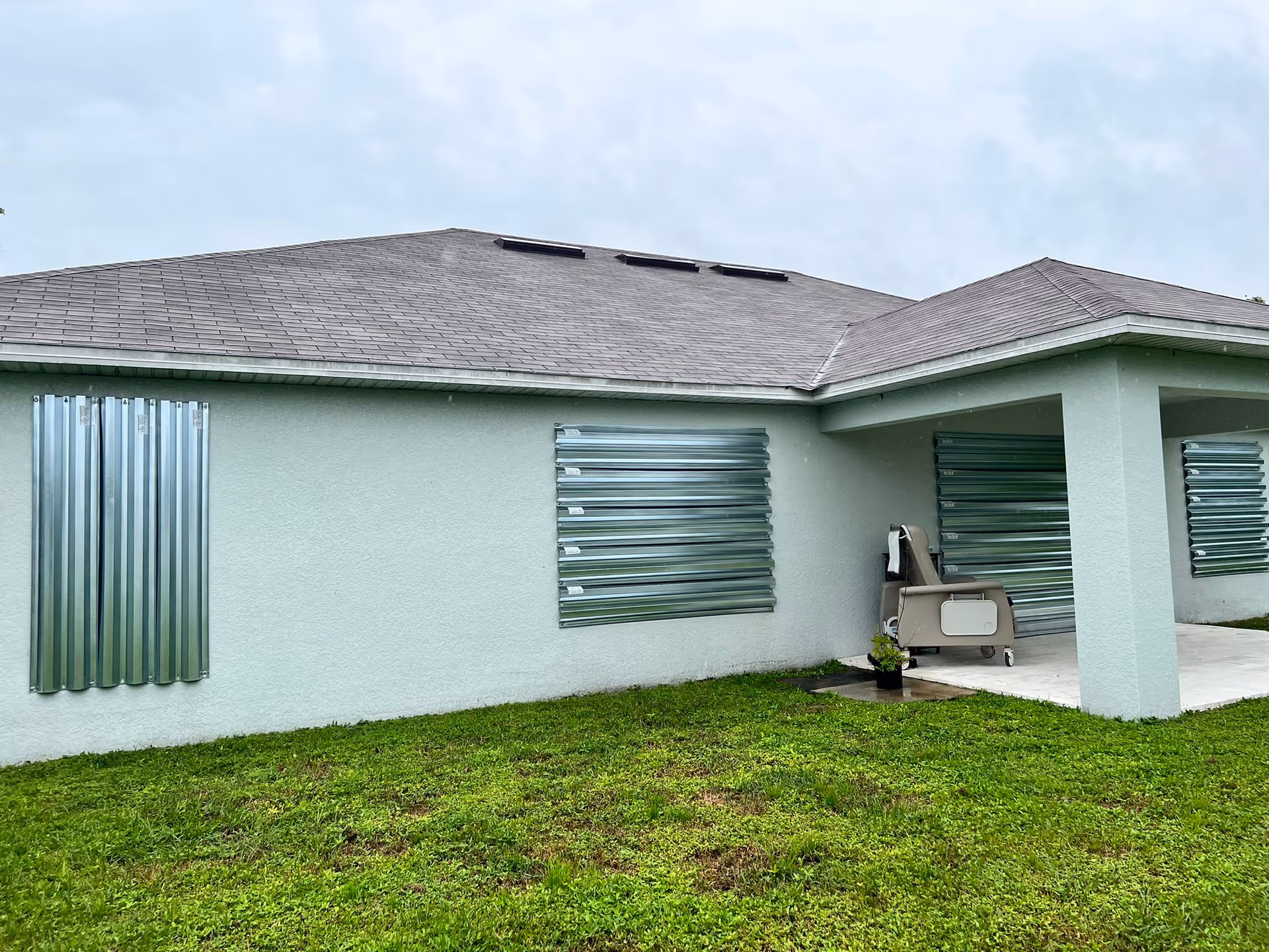 Single-story light-colored building exterior with metal hurricane shutters over windows and a covered patio on a grassy lawn under a cloudy sky.