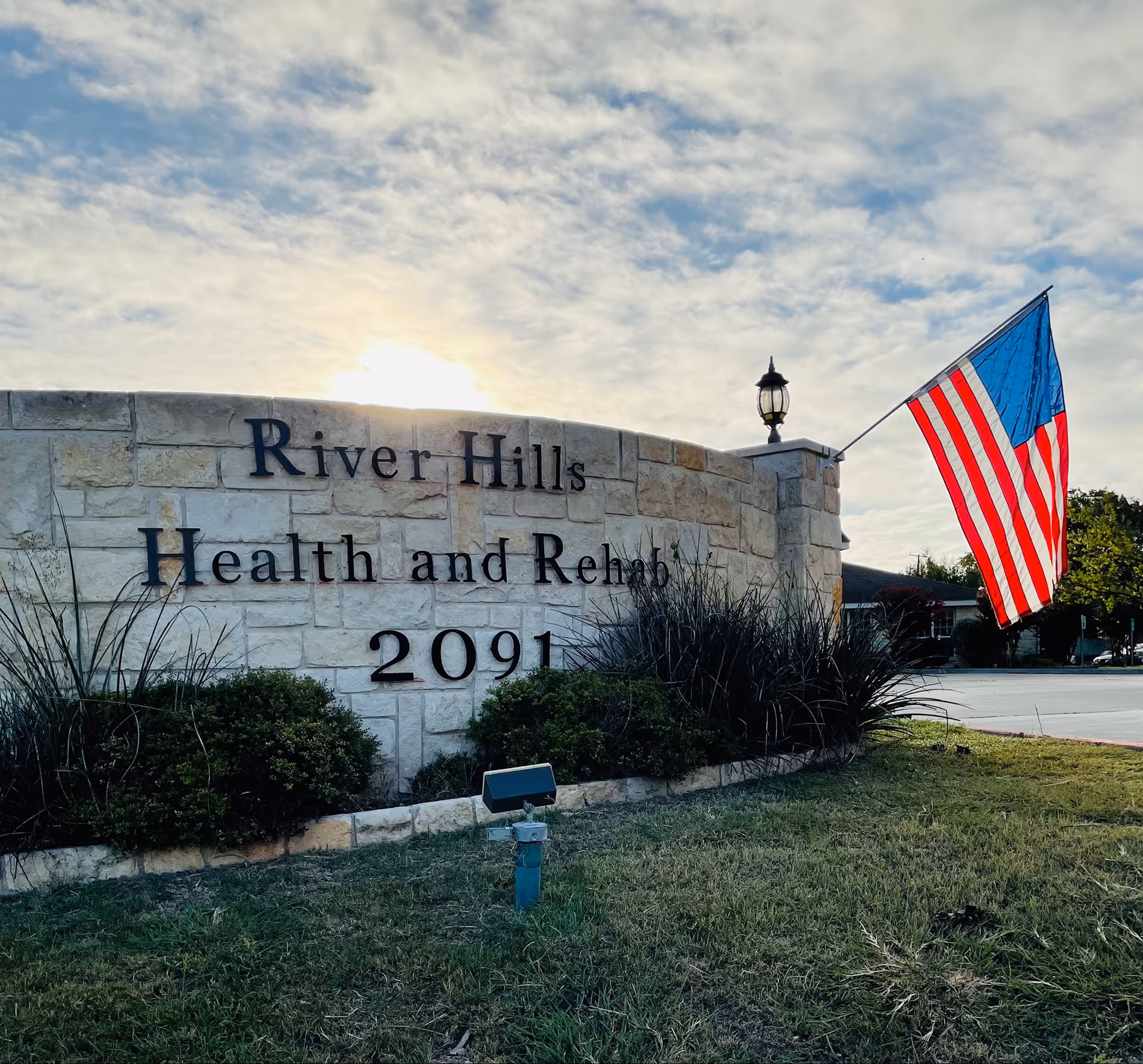 Stone sign for River Hills Health and Rehabilitation with the address number 2091, an American flag mounted on the right side, some bushes in front, and a partly cloudy sky in the background.