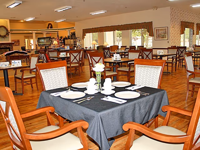 Spacious assisted-living dining room with a set table in the foreground and multiple tables and chairs throughout the room.