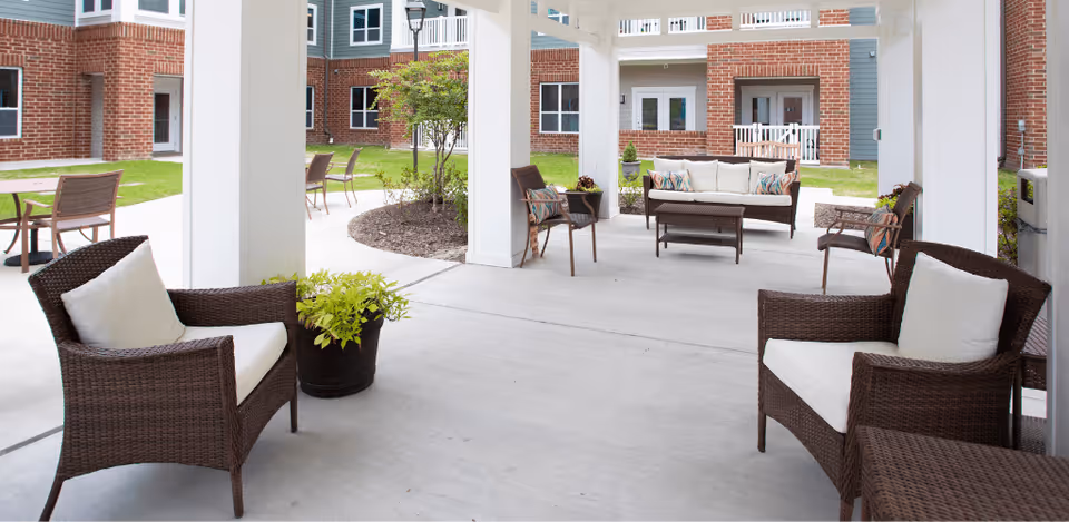 Outdoor covered seating area with wicker chairs and a sofa with cushions, surrounded by a garden and brick building in the background.