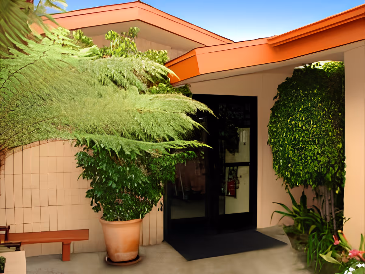 Entrance area of Acacia Villas featuring a glass door surrounded by beige walls and an orange roof overhang, with lush green plants in pots and landscaping around the doorway.
