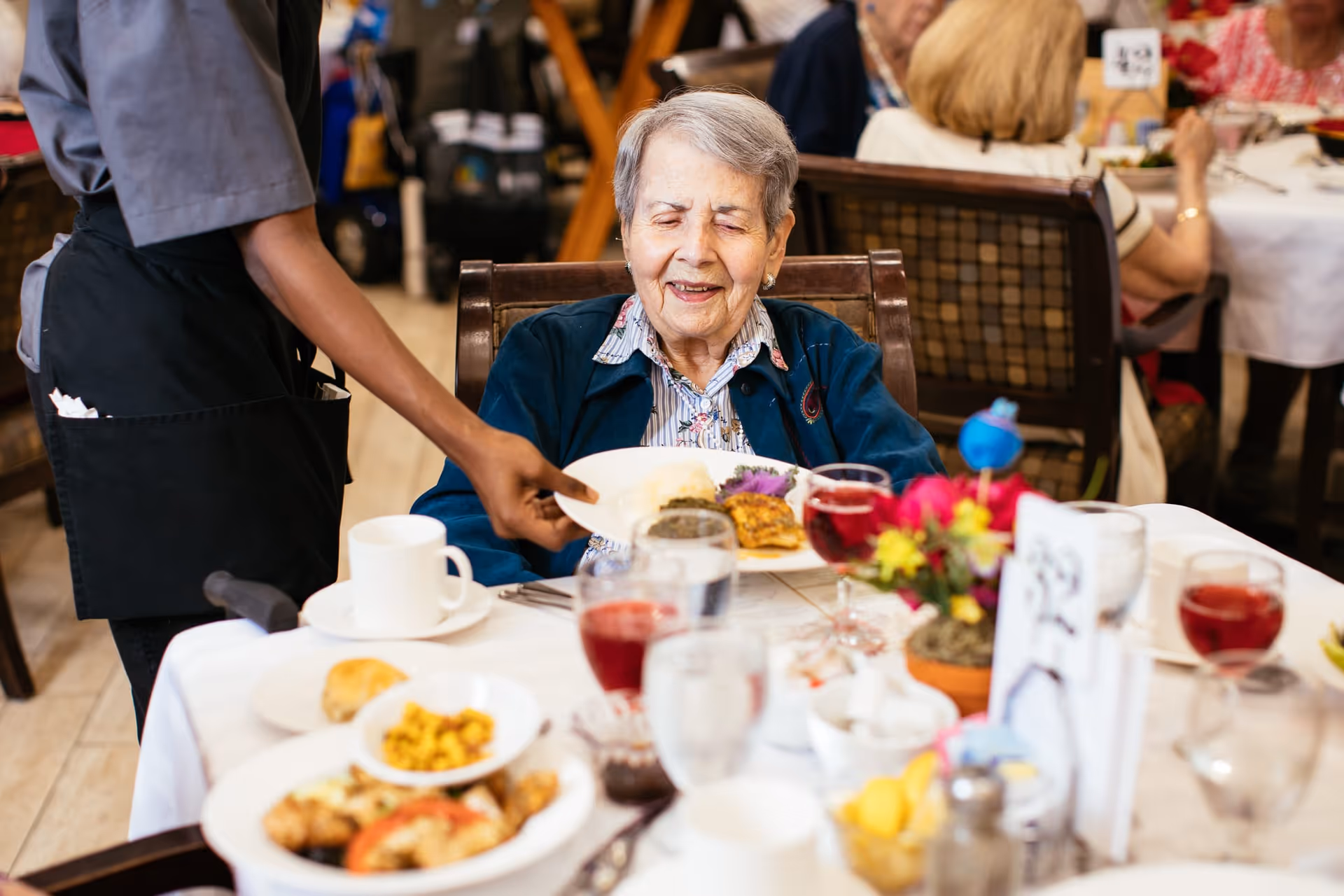 An elderly woman sitting at a dining table in a senior living facility, smiling as a server places a plate of food in front of her. The table is set with various dishes, drinks, and a small flower arrangement. Other seniors are visible dining in the background.