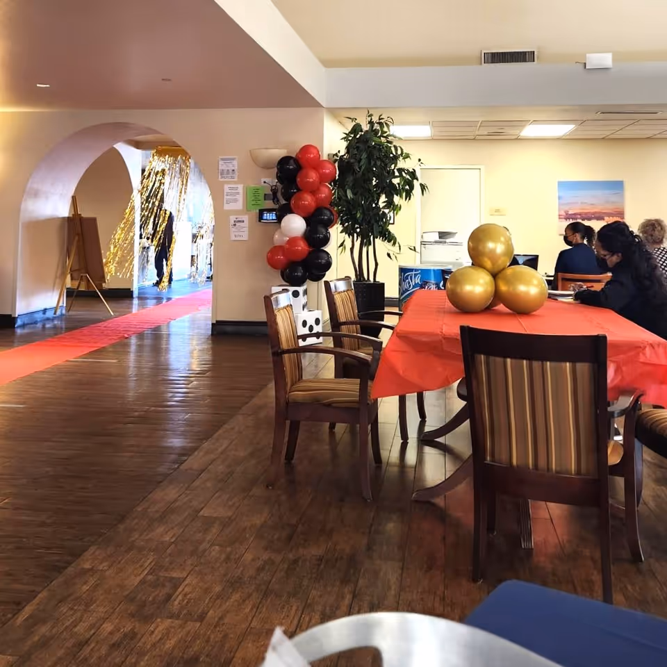 Interior view of a common area in an assisted living facility with wooden flooring, a table covered with a red tablecloth and decorated with gold balloons, several chairs around the table, a plant in the corner, and a cluster of red, black, and white balloons near an archway. People are seated at the table in the background.