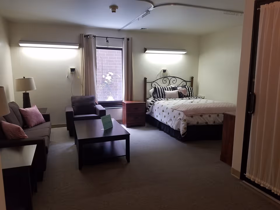 A cozy senior living room and bedroom area with a bed featuring a decorative headboard, patterned bedding, and pillows. The room also includes a sofa with pink cushions, an armchair, a coffee table, two nightstands, and a window with curtains. The walls are light-colored, and there are two wall-mounted lights above the bed and sofa.