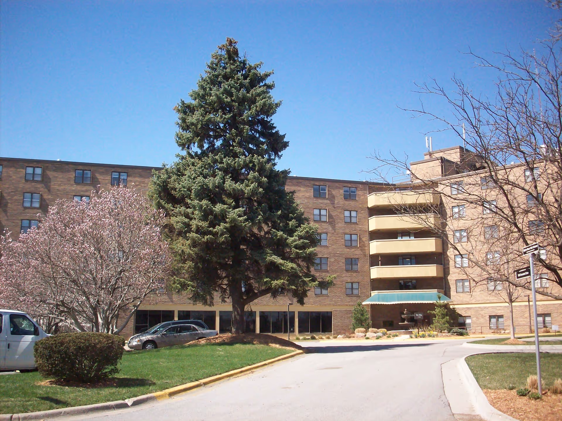 Exterior view of a multi-story senior living facility building with a brick facade, several windows, and balconies. In front of the building, there is a driveway, green lawn, a large evergreen tree, a blooming tree with pink flowers, and a few parked cars under a clear blue sky.