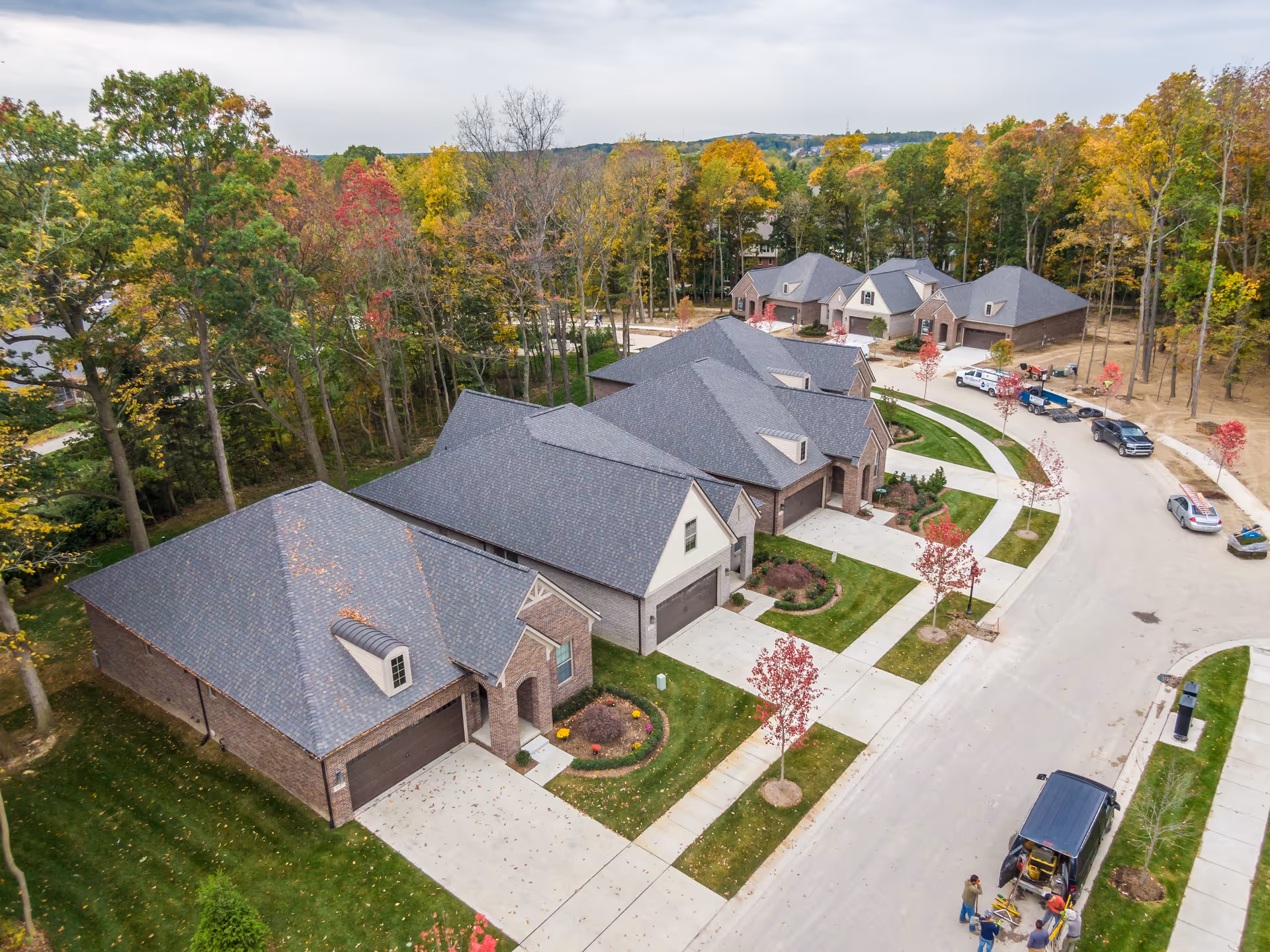 Aerial view of a residential neighborhood with several single-story brick houses featuring dark shingled roofs. The houses are lined along a curved street with neatly maintained lawns and young trees with autumn foliage. A few vehicles and people are visible on the street near the bottom right corner. The area is surrounded by a forest with trees showing fall colors.