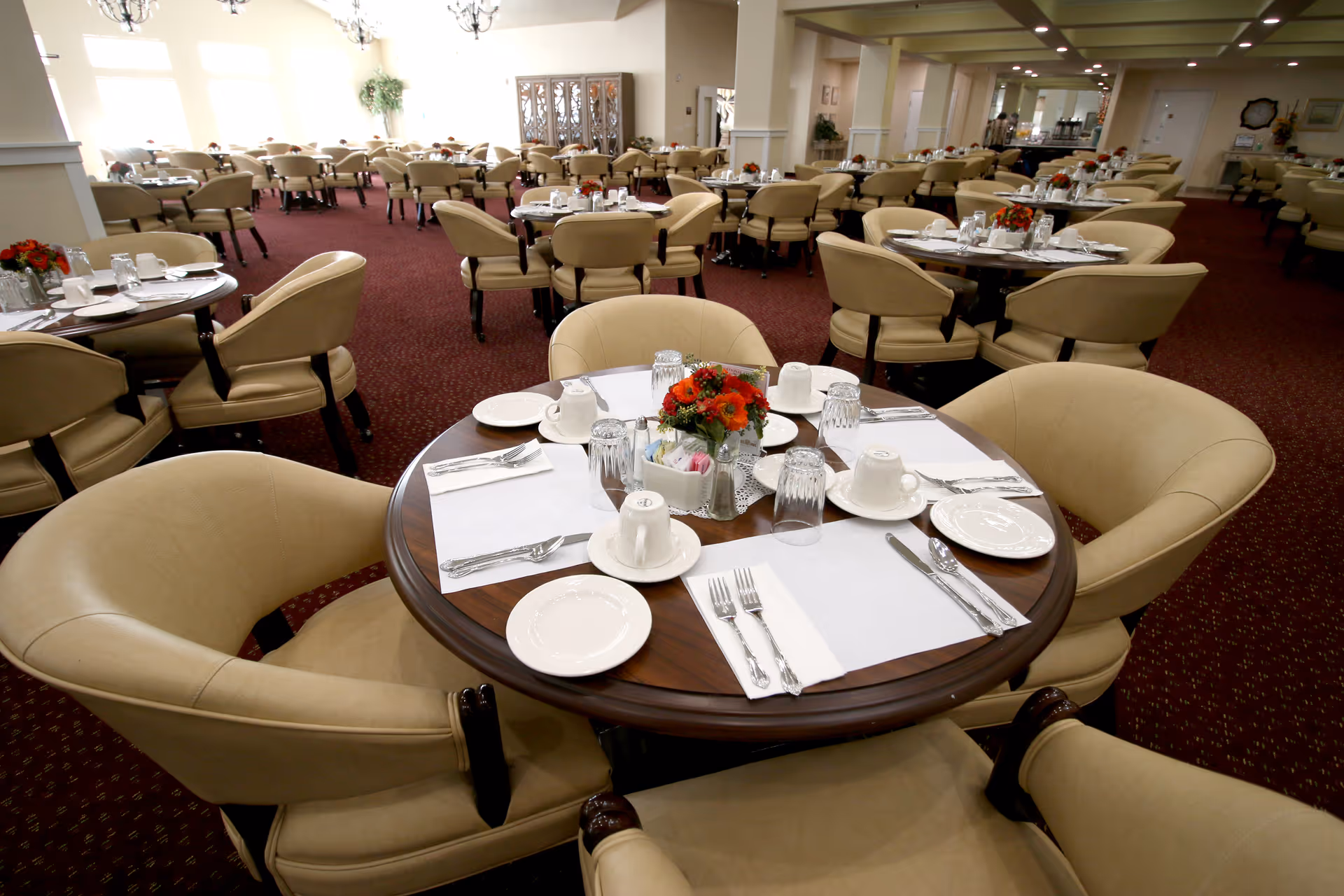 Spacious dining room with round tables set with place settings, floral centerpieces, and beige upholstered chairs.