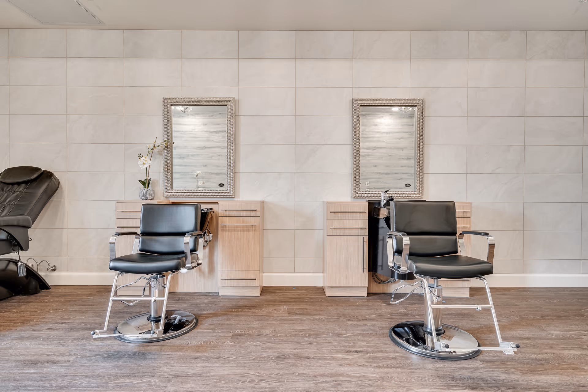 Two black salon chairs facing mirrors above light wood styling stations in a clean, tiled interior salon area.