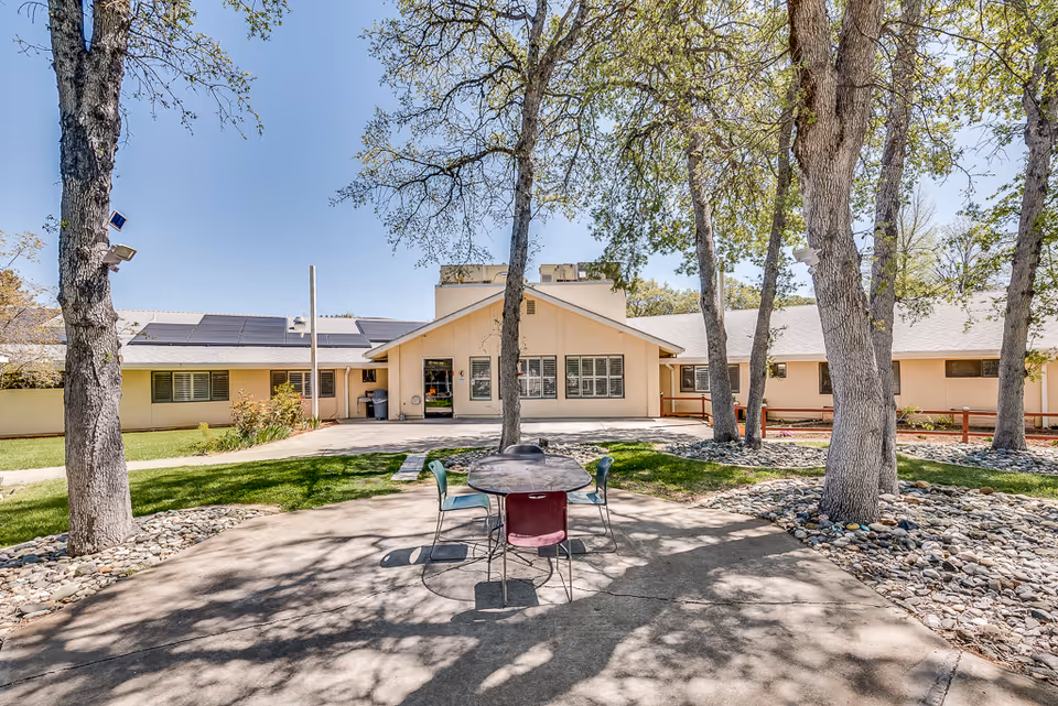 Outdoor courtyard area of Meadowood Nursing Center with a round table and four chairs on a concrete patio surrounded by trees and landscaping. The building is visible in the background under a clear blue sky.