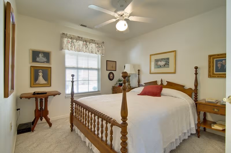 A cozy bedroom with a wooden four-poster bed covered in a white bedspread and a red pillow. The room has a window with a floral valance, a ceiling fan with a light, framed pictures on the walls, a wooden side table with a lamp, and a small wooden table near the window.