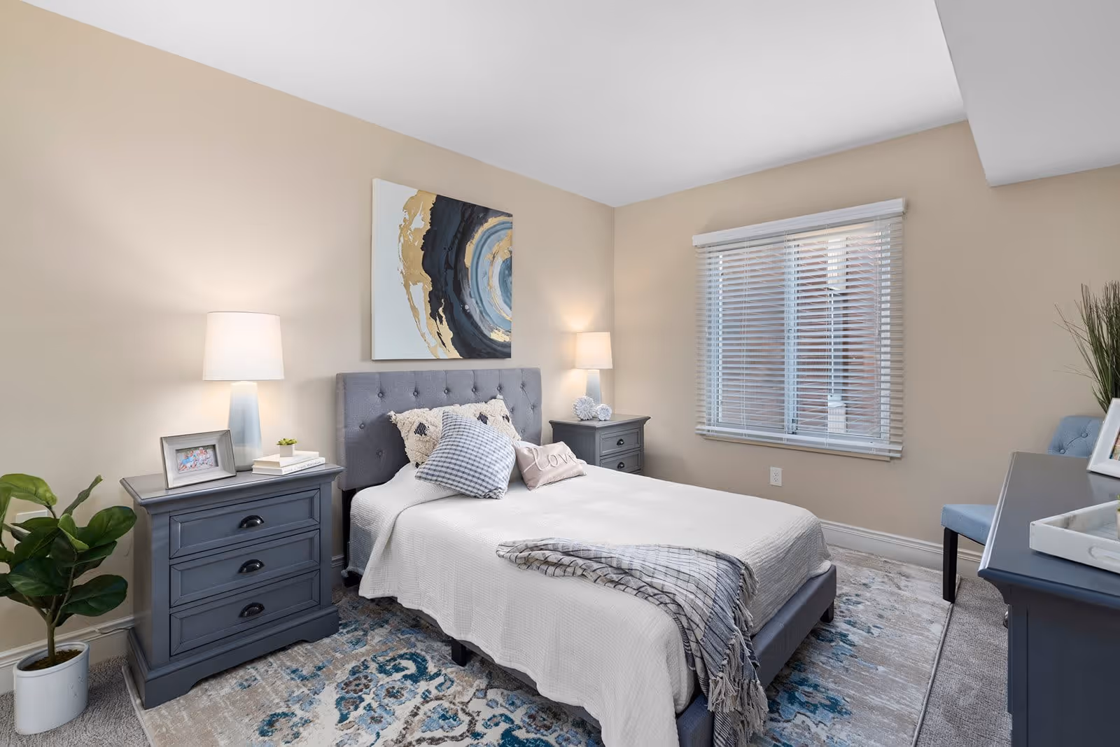 A neatly arranged bedroom with a gray upholstered bed featuring white bedding and decorative pillows. There are two matching gray nightstands on either side of the bed, each with a lamp. A large abstract painting hangs above the bed. A window with white blinds is on the right wall, and a blue patterned rug covers part of the carpeted floor. A potted plant is placed next to the left nightstand, and a blue chair is positioned near a dresser on the right side of the room.