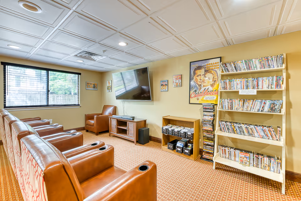 A cozy media room with a large flat-screen TV mounted on the wall, surrounded by four brown leather recliners and one armchair. There is a window with blinds on the left wall, and shelves filled with DVDs and VHS tapes on the right. The walls are painted beige and decorated with vintage movie posters, including one for Casablanca.