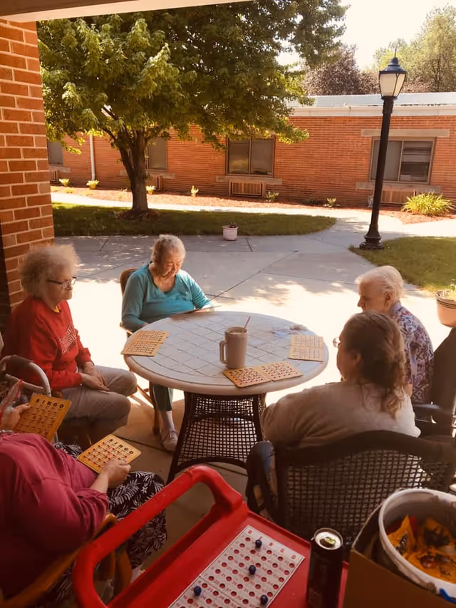 A group of elderly women sitting around a round outdoor table playing bingo. The table has bingo cards and a large cup with a straw. They are seated on a covered patio area with a brick wall and a tree in the background. A lamp post and some plants are visible in the courtyard beyond the patio.