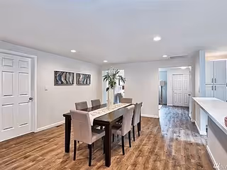 A modern dining area with a rectangular wooden table surrounded by six upholstered chairs. The room has light-colored walls, wood flooring, and recessed ceiling lights. There are two abstract paintings on the wall and a tall plant in a white vase on the table. The space opens into a hallway and kitchen area with white cabinetry.