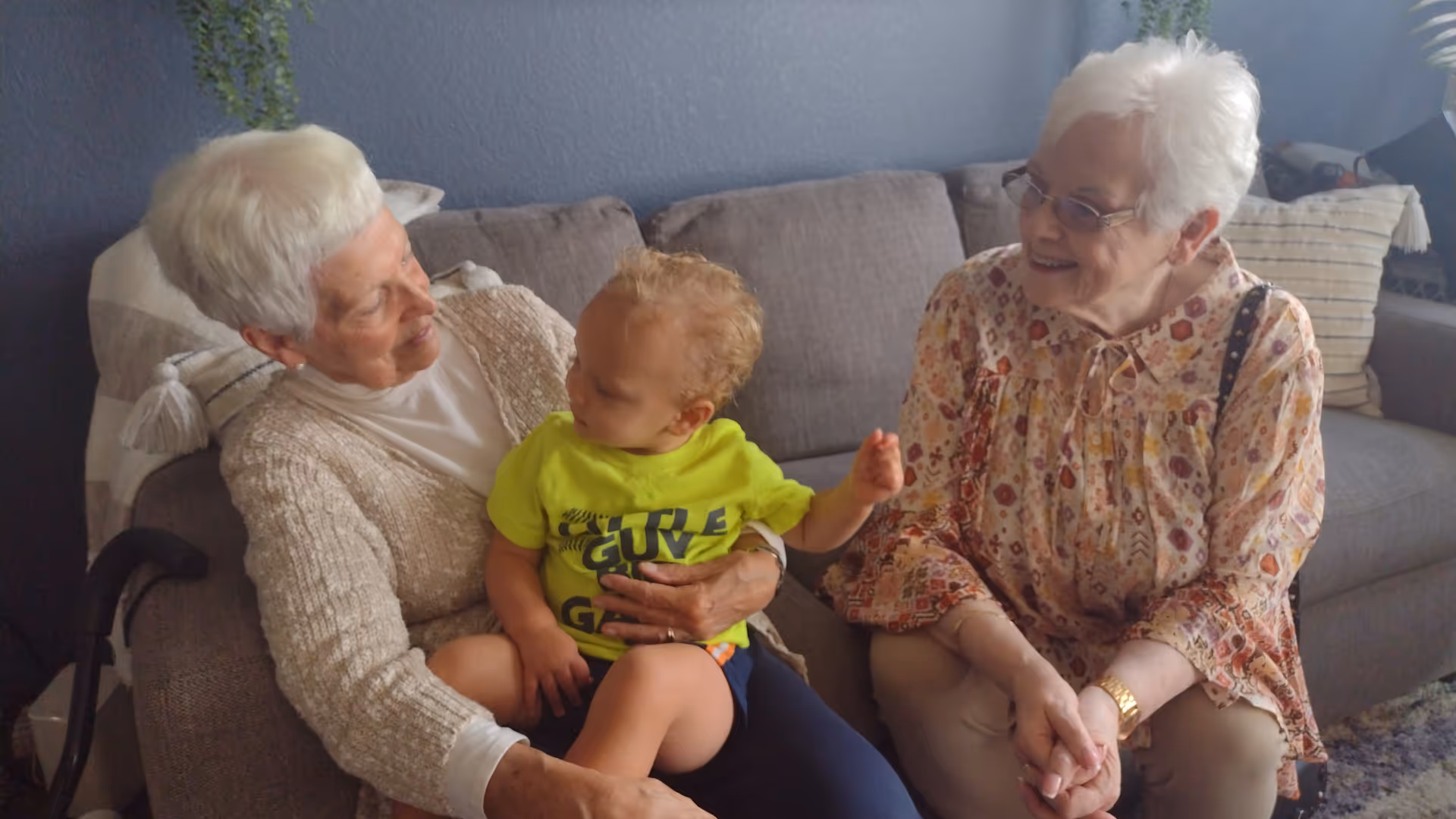Two elderly women smile at a toddler seated on a couch in a living room setting.