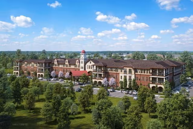 A large, multi-story senior living facility building with a clock tower and red roof accents, surrounded by trees and parking lots under a blue sky with scattered clouds.