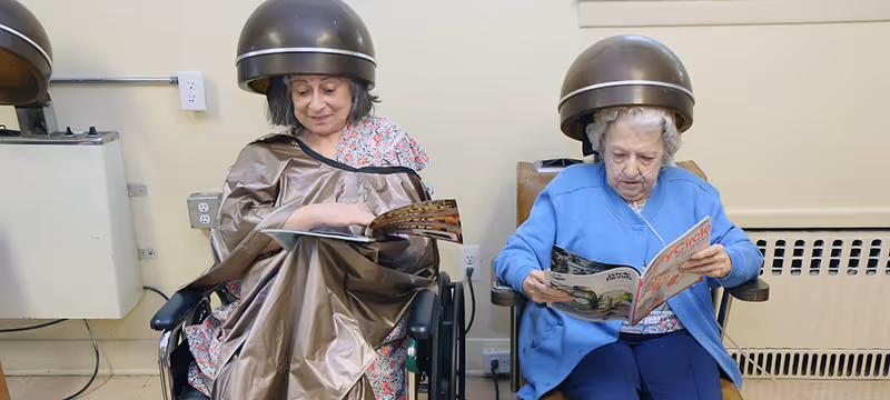 Two elderly women sitting under hair drying machines in a salon area of a senior living facility. One woman is wearing a brown salon cape and reading a magazine, while the other woman is dressed in a blue cardigan and also reading a magazine.