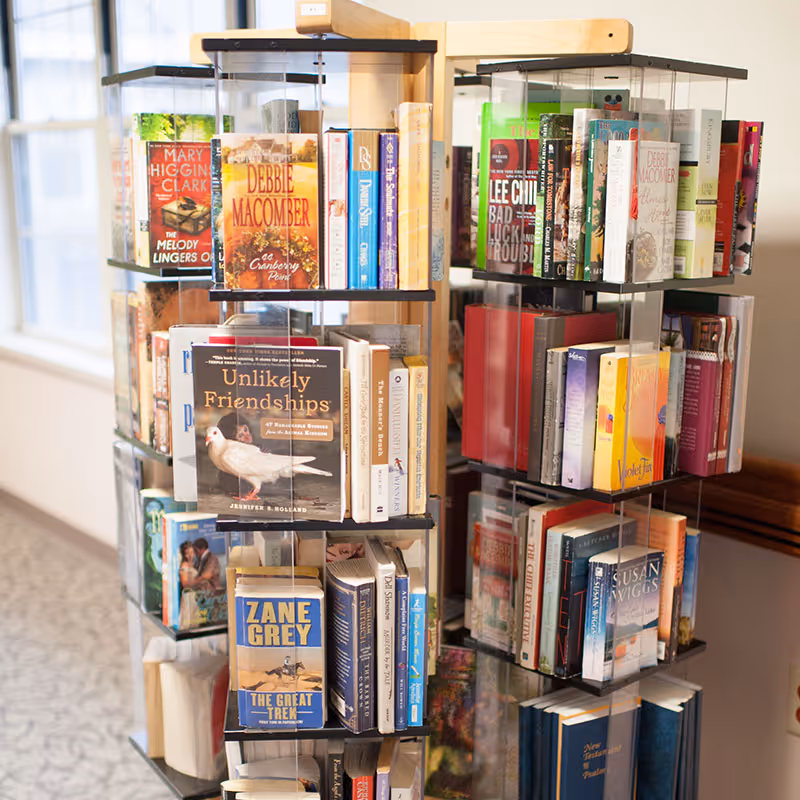 A rotating book display rack filled with various books including titles by Debbie Macomber, Mary Higgins Clark, and Zane Grey, situated in a well-lit indoor area with a window and carpeted floor visible in the background.