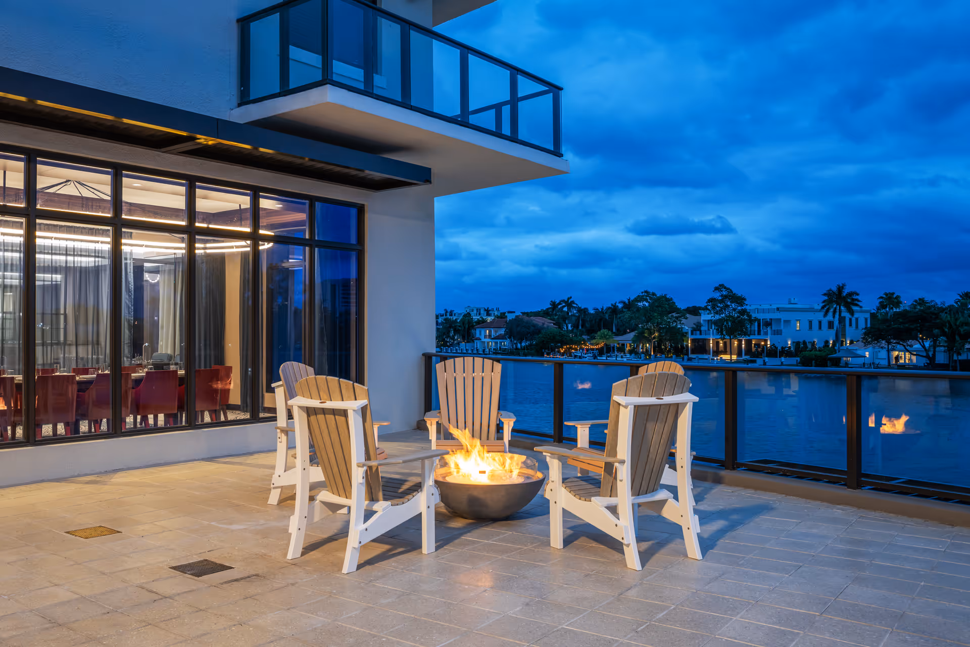 Outdoor patio area at dusk with four wooden Adirondack chairs arranged around a fire pit. The patio overlooks a body of water with houses and palm trees in the background under a cloudy blue sky. Large windows reveal an indoor dining area with red chairs and a modern light fixture.