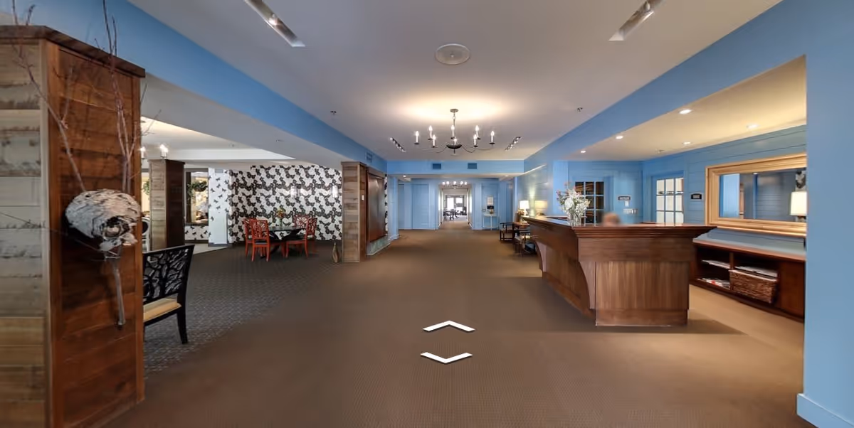 Interior view of a senior living facility lobby with a wooden reception desk on the right, blue walls, a chandelier hanging from the ceiling, and a seating area with red chairs and a table on the left. The space is well-lit with recessed lighting and has a carpeted floor.