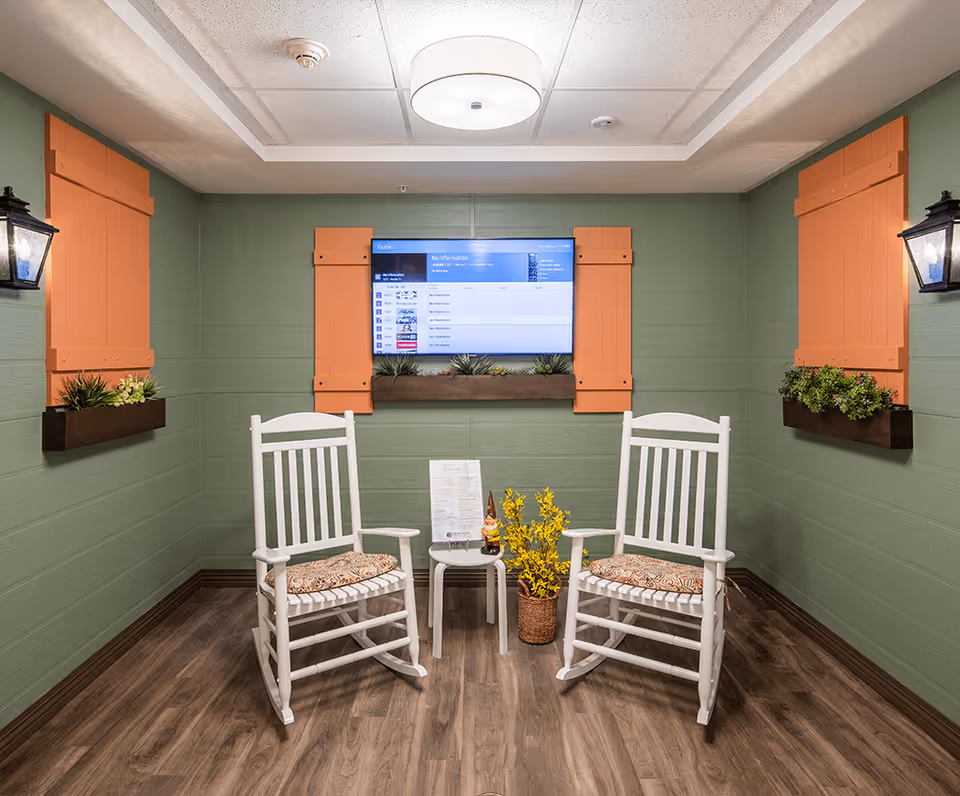A cozy seating area with two white wooden rocking chairs with patterned cushions, a small white side table between them holding a menu and a small decorative gnome, and a basket with yellow flowers on the floor. The walls are painted green with orange window shutters and flower boxes containing green plants. A flat-screen TV is mounted on the wall above the table, and two black lantern-style wall lights are on either side.