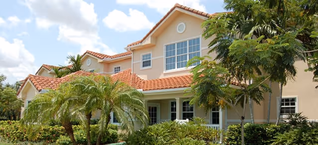 Exterior view of a two-story residential building with beige walls and a red tiled roof, surrounded by lush green palm trees and other tropical plants under a partly cloudy sky.
