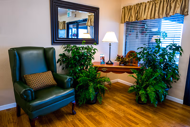 A cozy corner in a residential facility featuring a green leather armchair with a patterned cushion, a wooden side table with a lamp, decorative plants, and a large mirror on the wall reflecting part of the room and an exit sign.