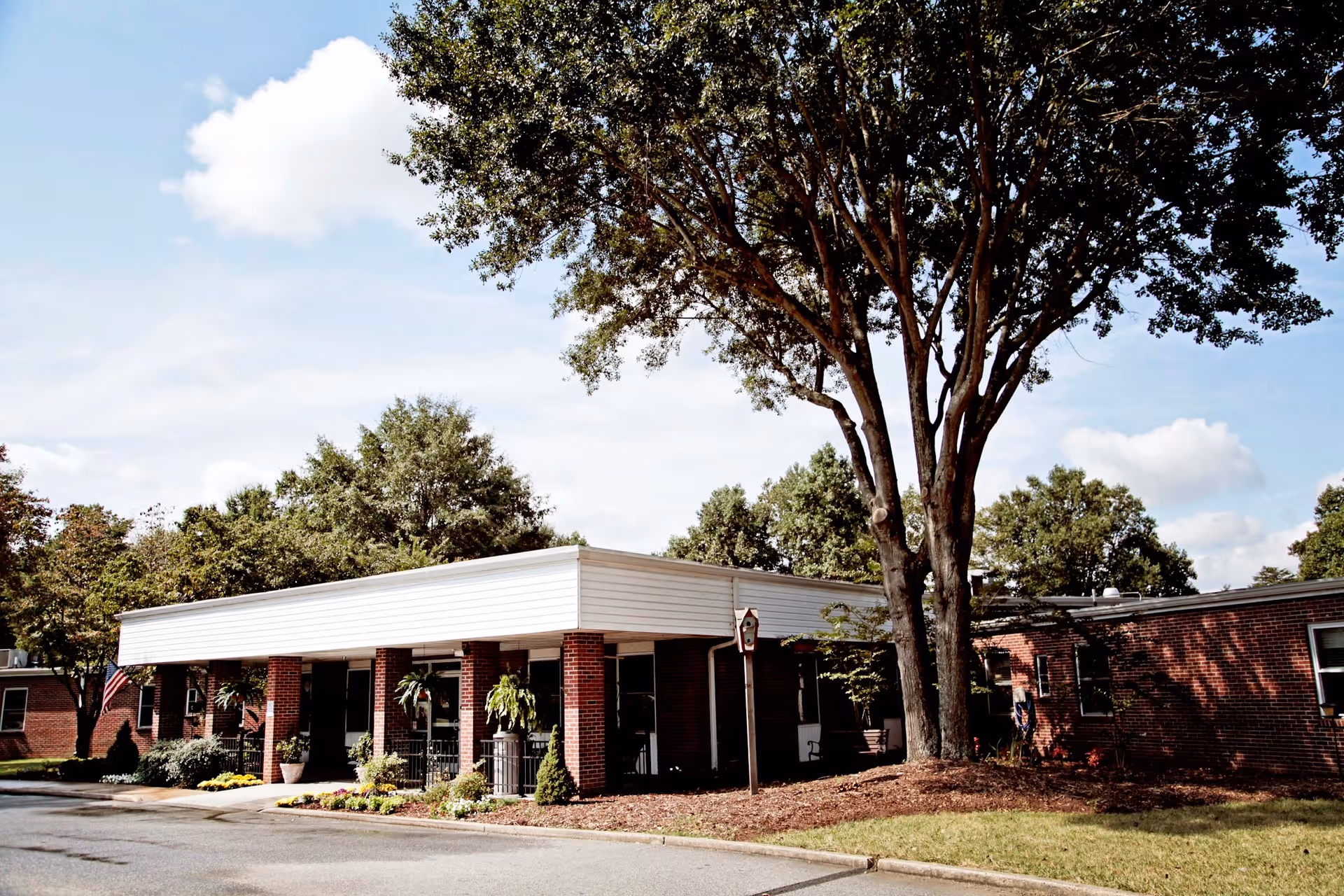 Exterior view of a single-story brick building with a white roof trim, surrounded by trees and landscaping under a partly cloudy sky.