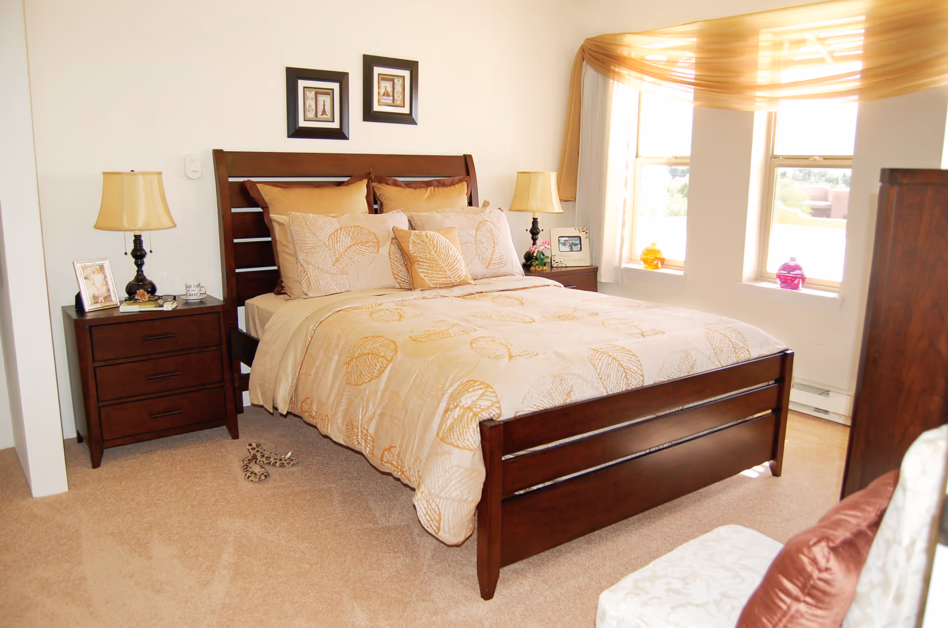 A bright bedroom with a wooden bed frame and beige bedding featuring a leaf pattern. There are two matching nightstands with lamps on either side of the bed. The room has beige carpet and two windows with sheer golden curtains letting in natural light. A pair of leopard print slippers is on the floor near the bed.