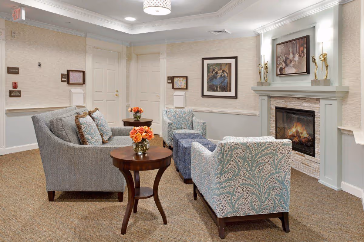 A cozy sitting area in a senior living facility with a gray sofa and two patterned armchairs arranged around a fireplace. There are two wooden side tables with vases of orange roses, framed artwork on the walls, and soft lighting from ceiling fixtures and wall sconces.