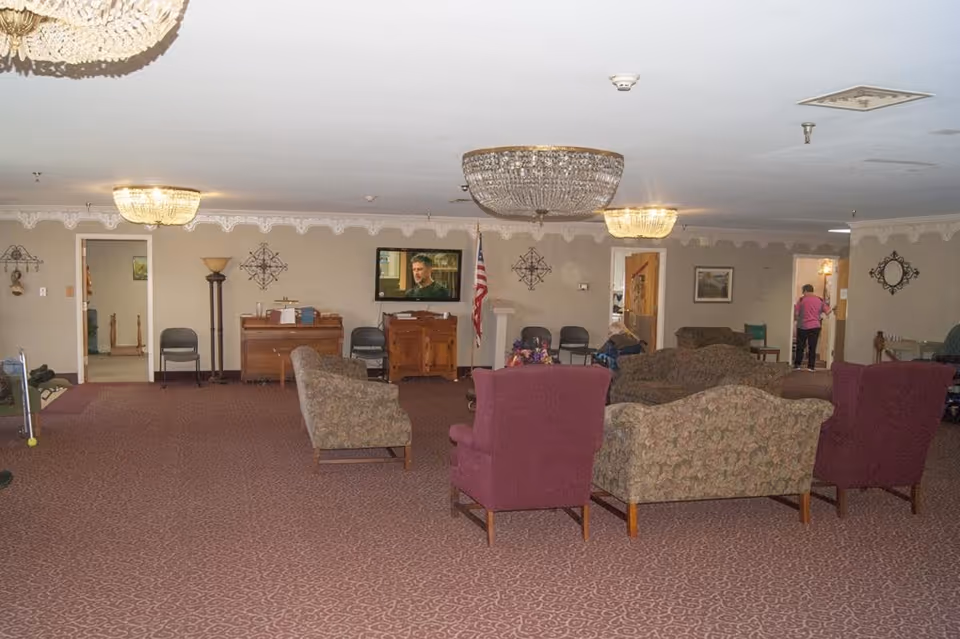 A spacious living room area in a senior living facility with patterned carpet and multiple upholstered chairs and sofas arranged facing a wall-mounted television. The room is decorated with chandeliers, wall art, and an American flag near the TV. There are a few people visible in the background near doorways.