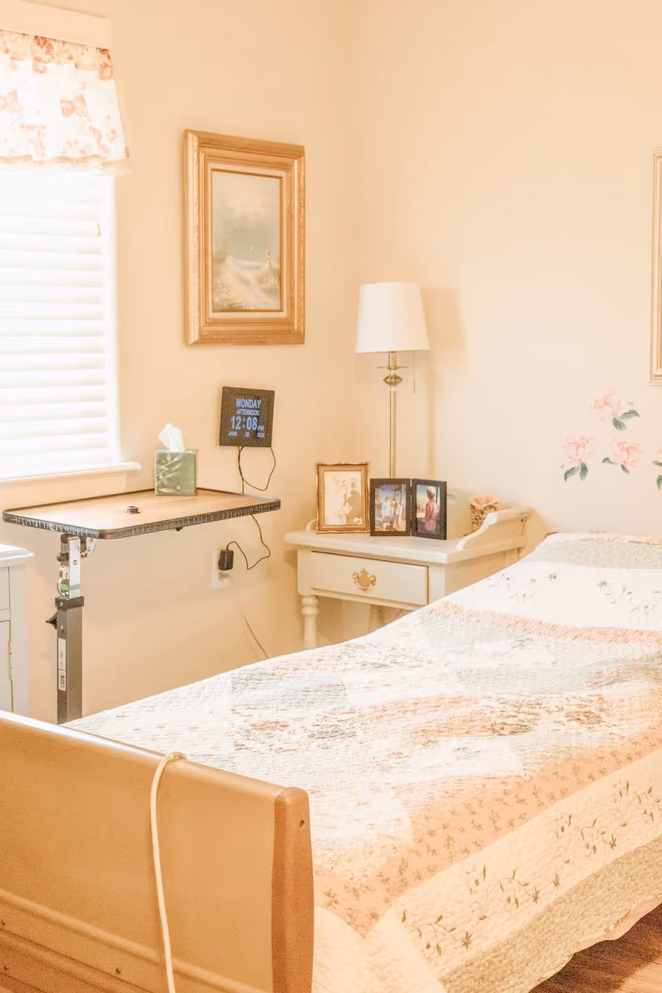 A cozy bedroom with a single bed covered in a patchwork quilt. Next to the bed is a small white nightstand with framed photos and a lamp. A small adjustable overbed table holds a tissue box. A digital clock on the wall shows the day and time. The room has soft beige walls and a window with floral curtains.