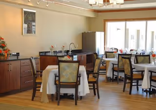 Dining room with round tables covered in white tablecloths, wooden chairs, a buffet counter and large windows letting in natural light.