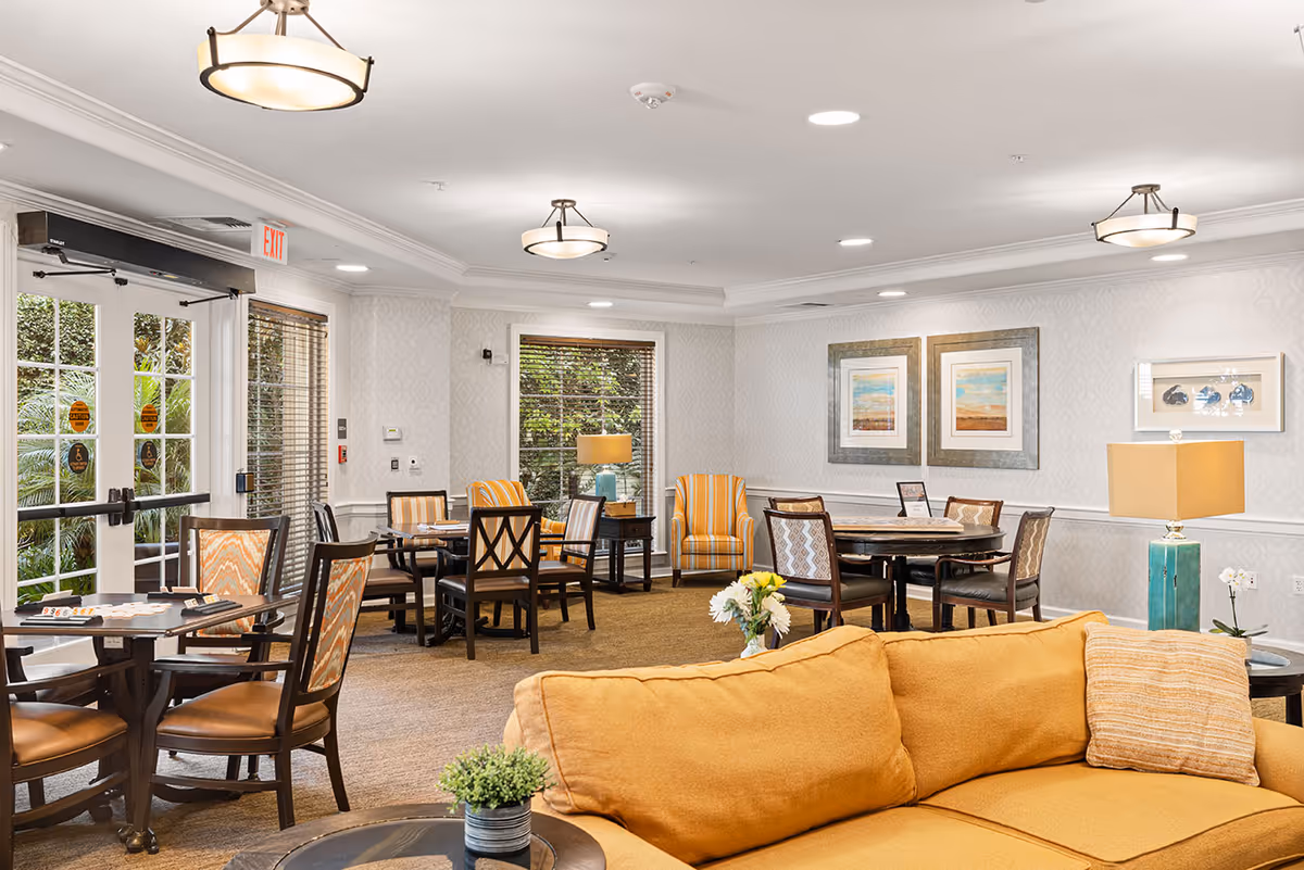 Bright communal lounge and dining area with round tables, chairs, framed art, and a yellow sofa.