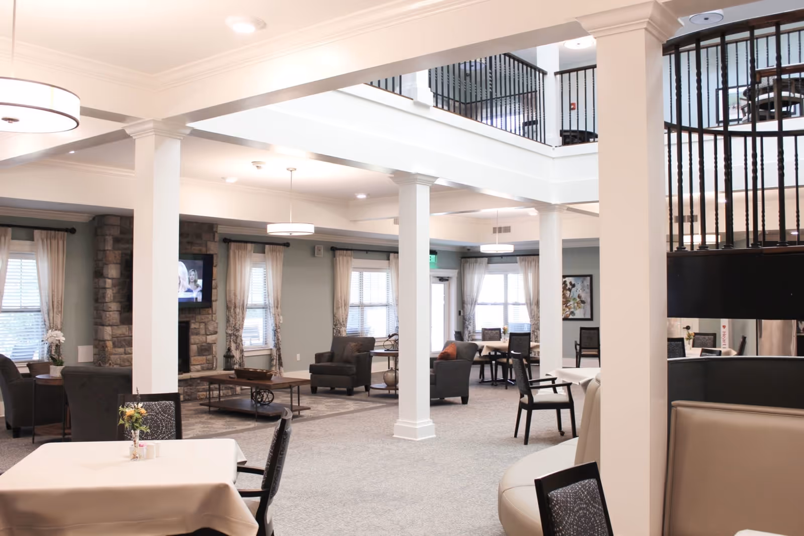 A spacious and well-lit senior living common area with multiple seating arrangements including armchairs and tables. The room features large windows with curtains, white pillars, a stone fireplace with a mounted TV, and modern light fixtures. The upper level with black railings is visible, overlooking the area below.