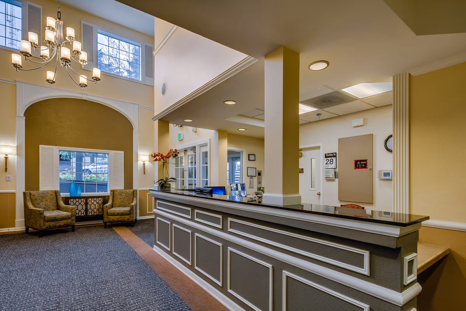 Reception area of Fred Lind Manor featuring a long counter with a black countertop, two patterned armchairs beside a window with white shutters, a chandelier hanging from the ceiling, and a carpeted floor.