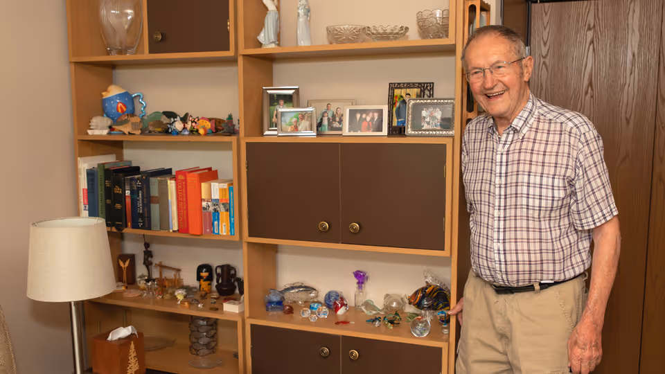 An elderly man wearing glasses, a plaid short-sleeve shirt, and beige pants stands smiling next to a wooden shelving unit filled with books, framed photos, decorative glass items, and various knick-knacks. A floor lamp and tissue box are visible to the left.