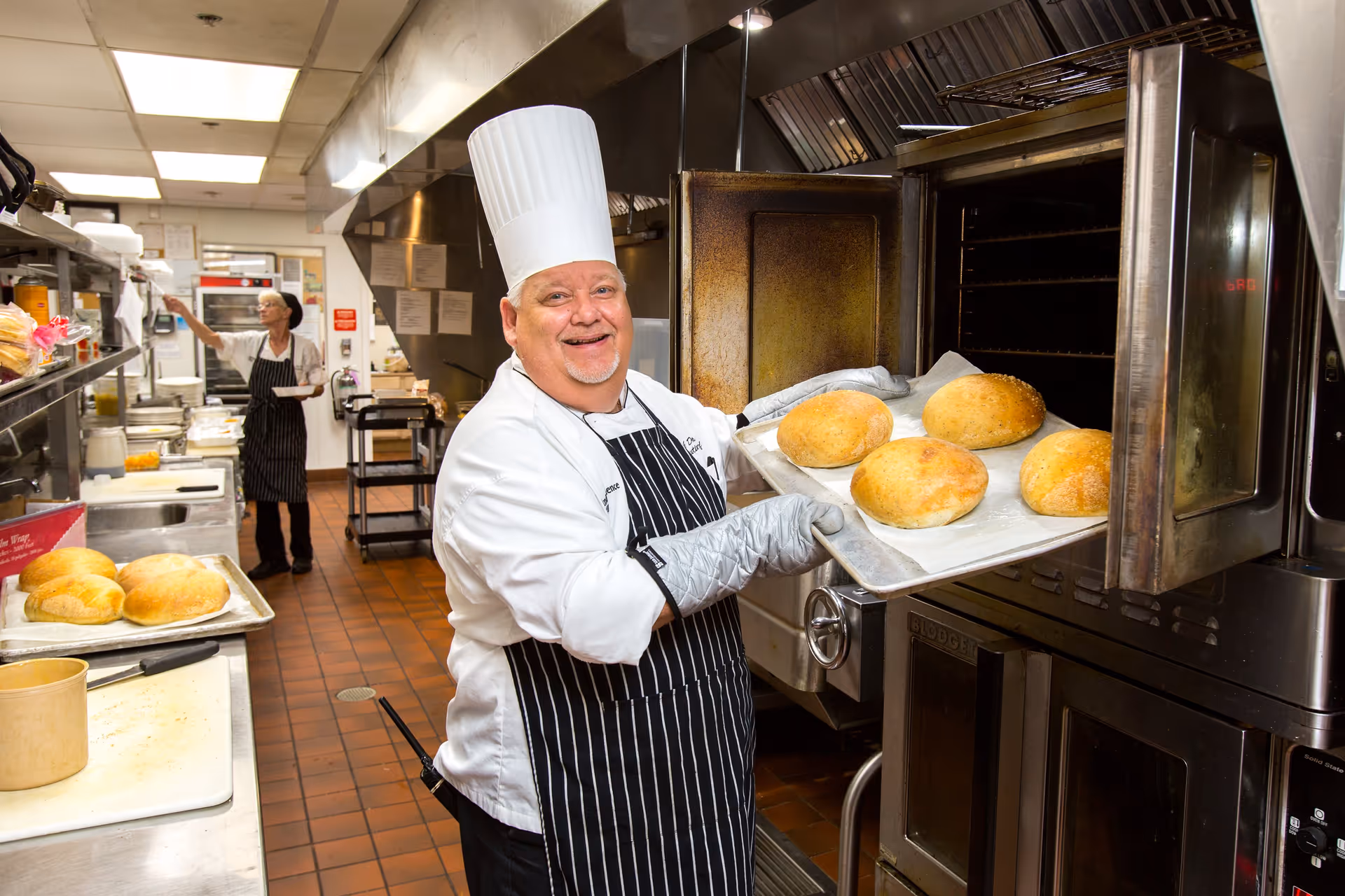 A chef wearing a white chef's hat, white coat, and striped apron is smiling while holding a baking tray with freshly baked bread rolls coming out of an industrial oven in a commercial kitchen. In the background, another kitchen staff member is reaching for something on a shelf.
