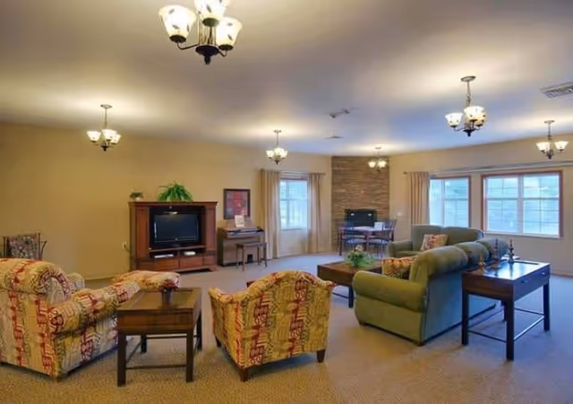 A spacious senior living common area with multiple seating options including patterned armchairs and green sofas arranged around wooden coffee tables. The room features beige walls, carpeted floors, several ceiling light fixtures, large windows with curtains, a TV on a wooden stand, a piano, and a small dining table near a stone fireplace.