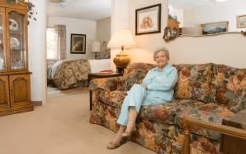 An elderly woman sitting on a floral patterned couch in a living room area of an assisted living facility. Behind her, there is a bedroom with a bed covered in a floral bedspread. The room is warmly lit with a table lamp on a side table next to the couch. The walls are decorated with framed pictures and a mirror. A wooden china cabinet is visible on the left side of the image.