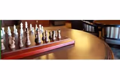 Wooden table with a chessboard and chess pieces in a communal seating area with upholstered chairs in the background.