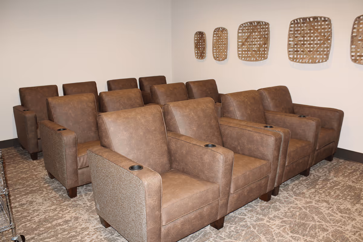 A small theater room with two rows of brown leather recliner chairs, each with a cup holder in the armrest. The walls are light-colored and decorated with woven basket-style wall art. The floor is carpeted with a patterned design.