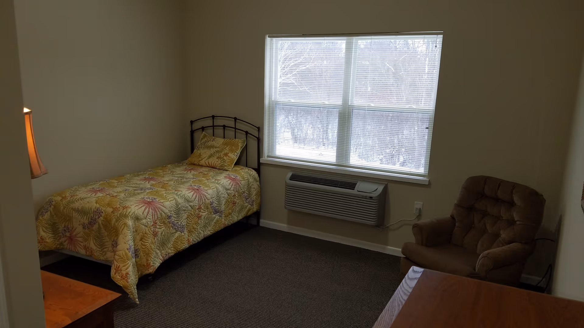 A small bedroom with a single bed covered with a floral patterned bedspread and a matching pillow. There is a window with blinds above a wall-mounted air conditioning unit. A brown cushioned armchair is placed near the window, and a wooden desk is partially visible in the foreground. The walls are plain and light-colored.
