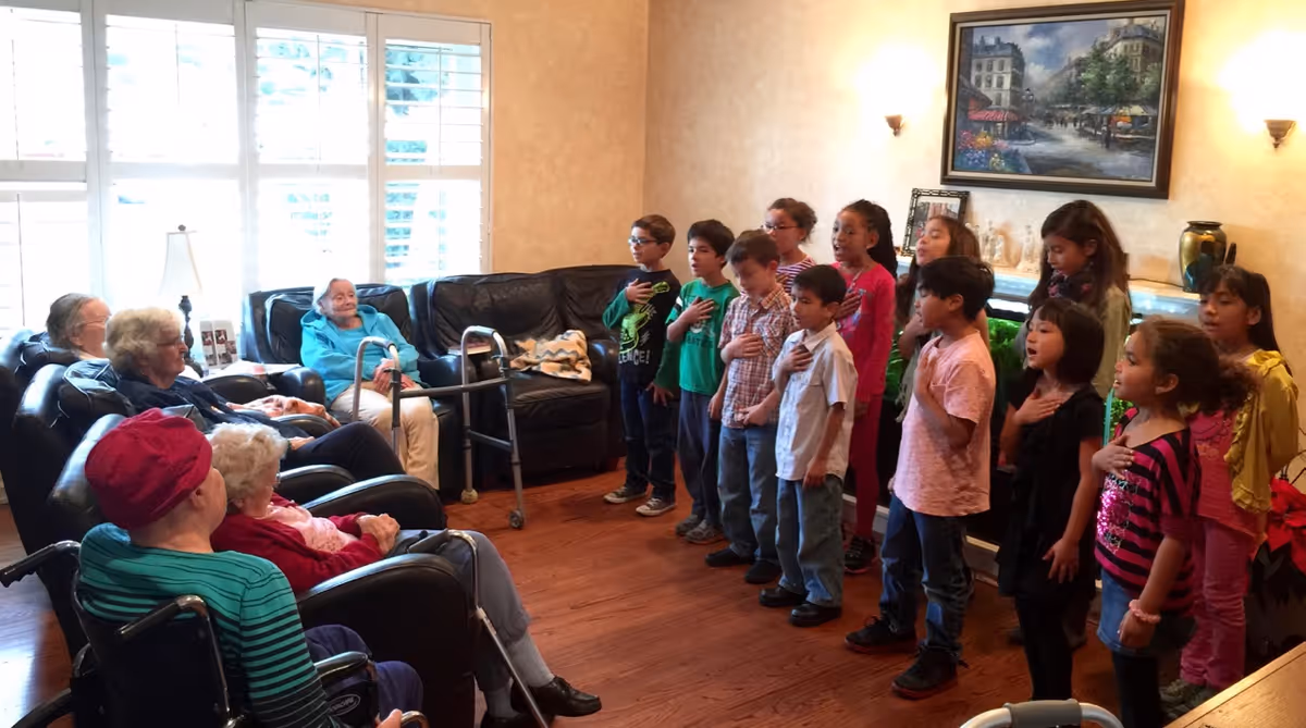 A group of elderly people seated in a living room watching a group of children standing and singing with their right hands over their hearts. The room has wooden floors, a large window with white shutters, a black leather couch, and a painting on the wall above a fireplace.