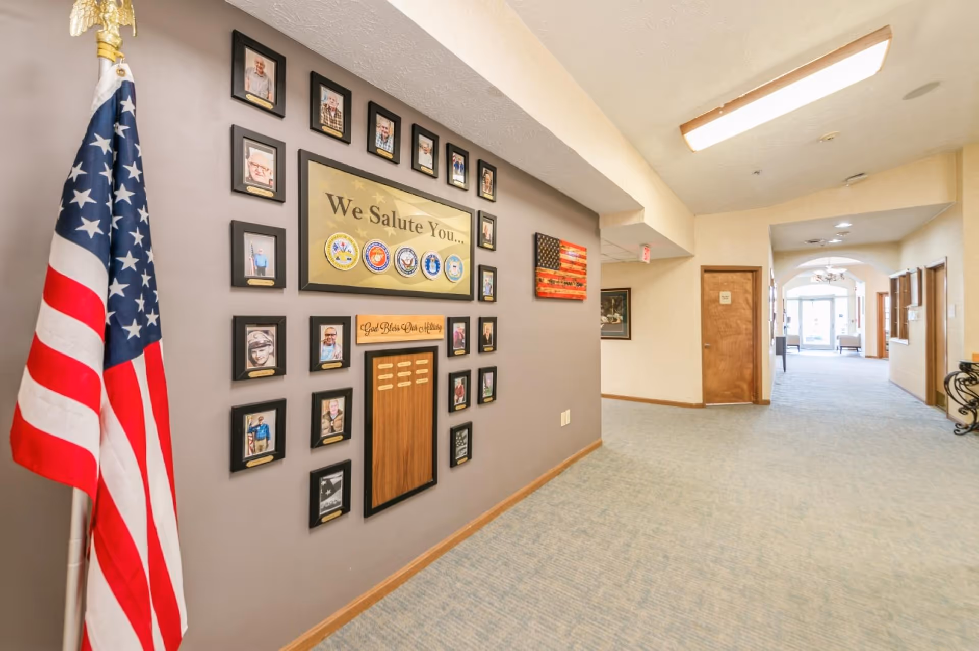 Interior hallway of a senior living facility with a patriotic display on the wall featuring framed photos of veterans, military emblems, and an American flag on a stand. The hallway is carpeted and leads to a glass door entrance with natural light.