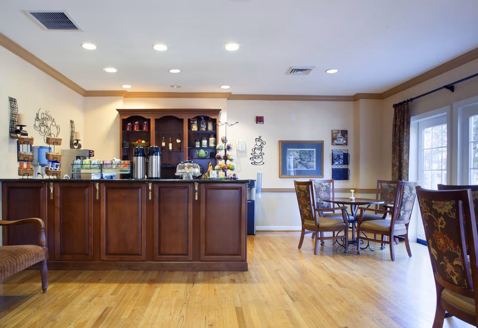 A bright common-area café with a wooden service counter, coffee dispensers, display cases and small dining tables with chairs by large windows.