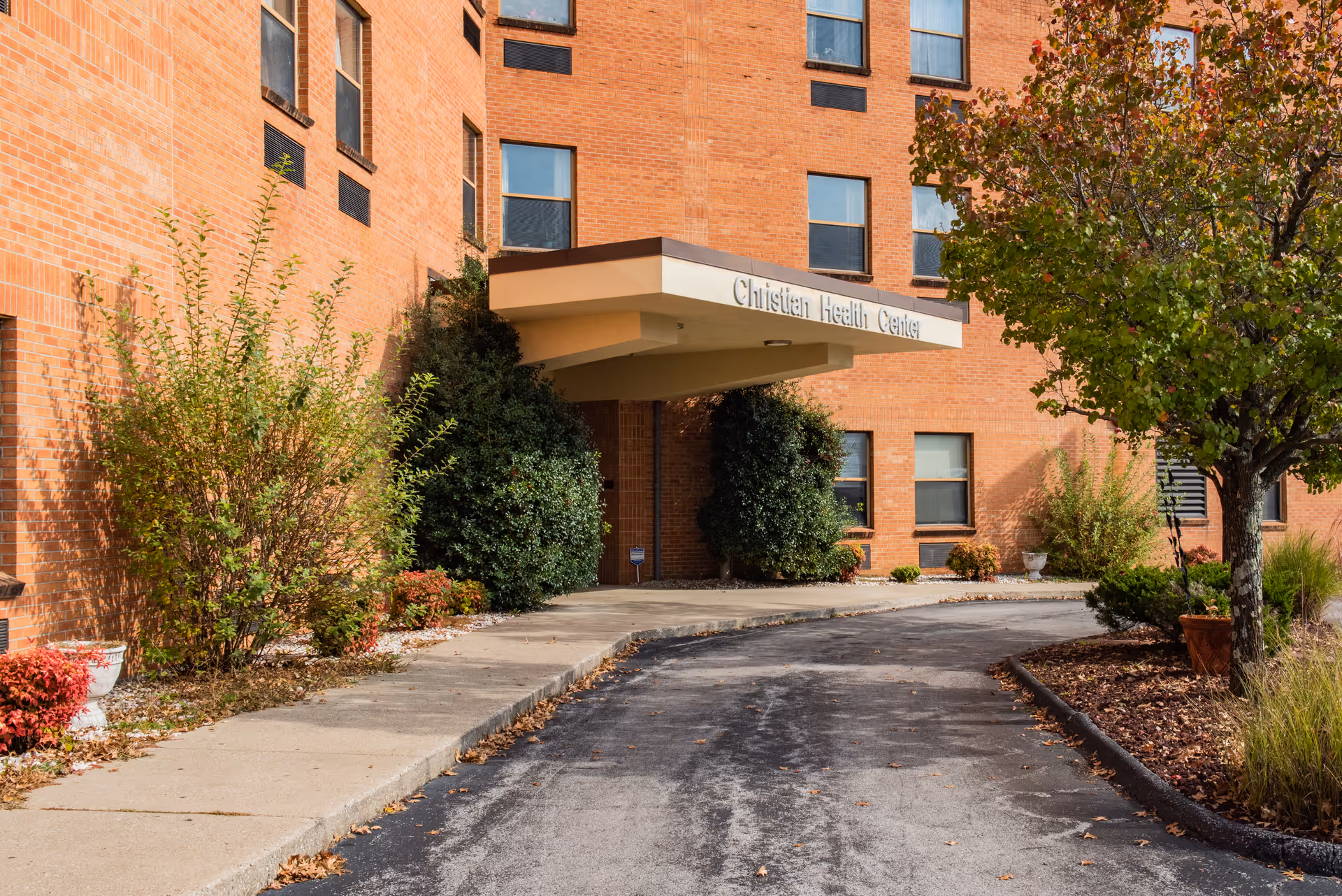 Front entrance and drive of the Christian Health Center brick building with a canopy, shrubs, and a tree.
