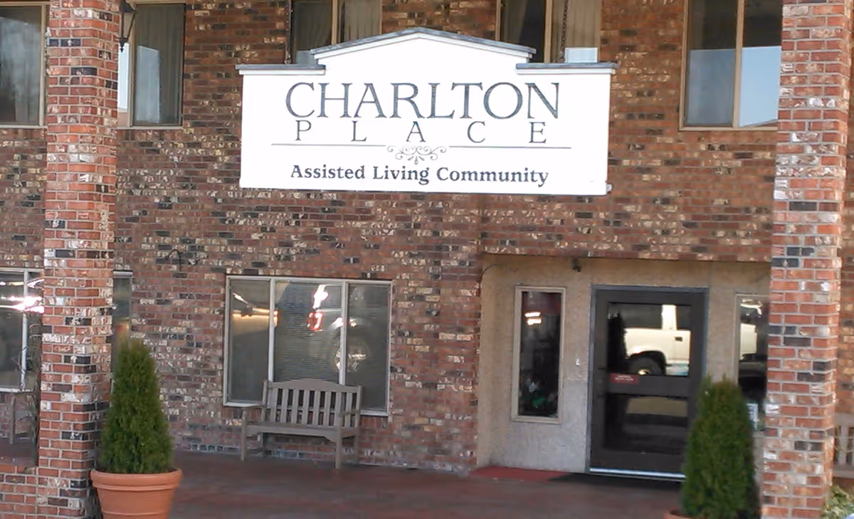 Entrance of Charlton Place Assisted Living Community with a brick exterior, a bench under a window, two potted plants, and a glass door.